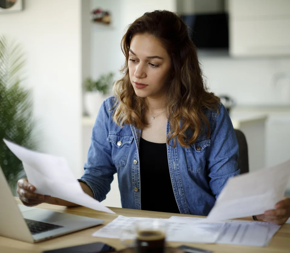 Woman with brown hair and wearing a blue long sleeve shirt, sat at a table, looking at paperwork